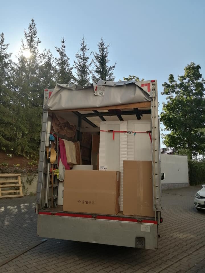 Otwarta przestrzeń ładunkowa wypełniona kartonami i meblami w trakcie załadunku, widok z tyłu pojazdu na tle drzew i słonecznego nieba.