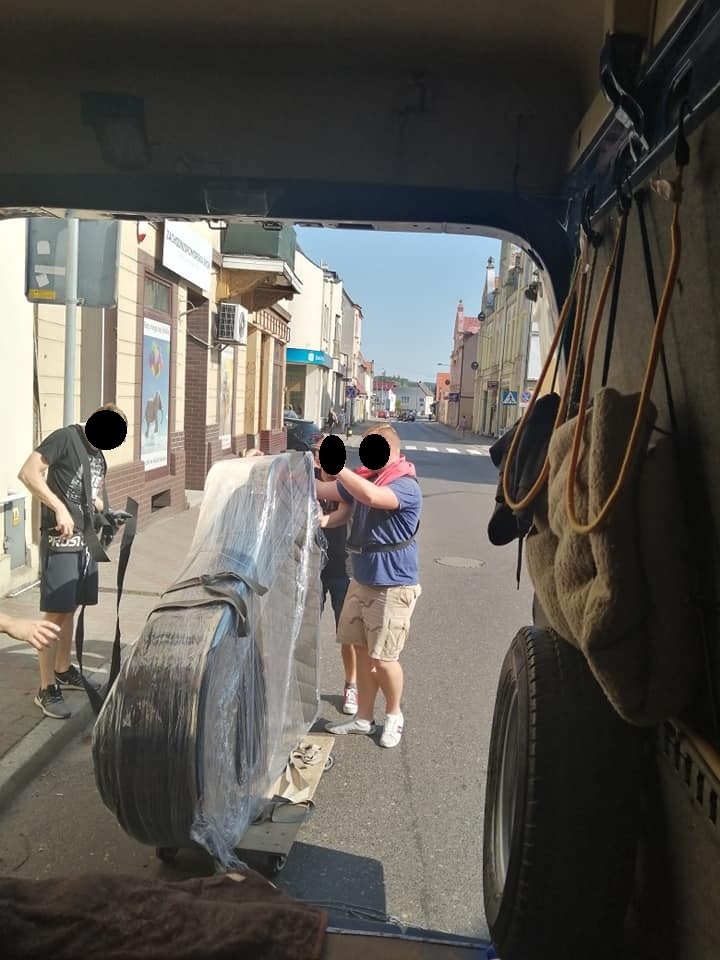 Trzech mężczyzn przenosi pianino owinięte folią z wózkiem do wnętrza furgonetki na tle ulicy miejskiej.