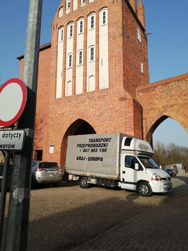 Biały samochód dostawczy z napisem 'Transport Przeprowadzki' i numerem telefonu na tle ceglanej bramy miejskiej i znaku drogowego.