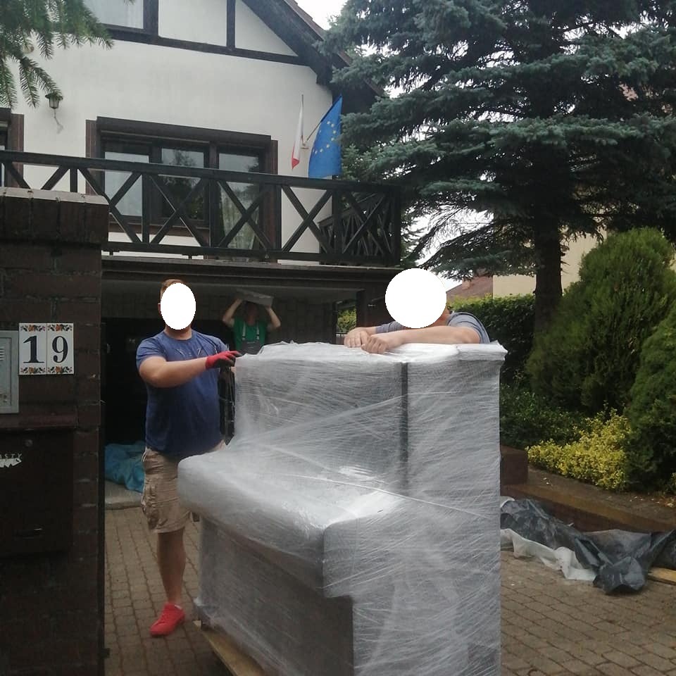Dwóch mężczyzn w trakcie transportu owiniętego folią fortepianu na palecie przed domem z numerem 19, trzecia osoba podtrzymuje element konstrukcyjny nad głową.
