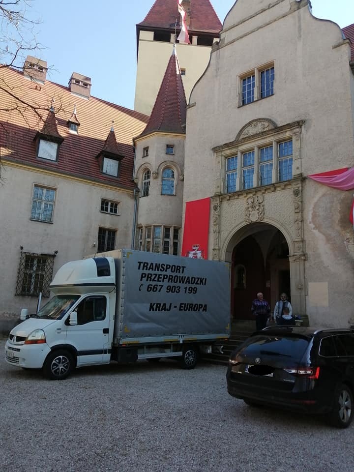 Biała furgonetka z szarym plandekowym nadwoziem z napisem 'TRANSPORT PRZEPROWADZKI (667 903 199) KRAJ - EUROPA' zaparkowana na żwirowym podłożu przed wejściem do zabytkowego zamku z czerwonymi...