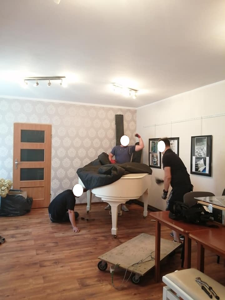 Trzech mężczyzn przygotowuje biały fortepian do transportu, owinięty częściowo kocem ochronnym, na drewnianej podłodze, z wózkiem transportowym w pobliżu.