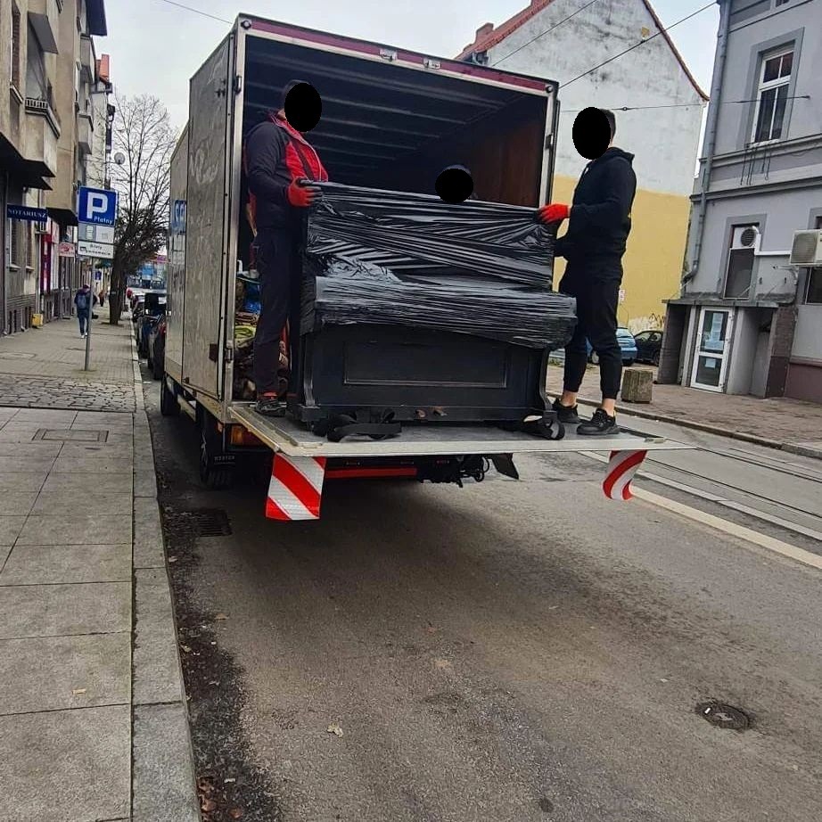 Dwóch mężczyzn w odblaskowych kamizelkach i czerwonych rękawicach ostrożnie przenosi pianino owinięte czarną folią z windy załadowczej samochodu dostawczego na ulicę w miejskiej scenerii.