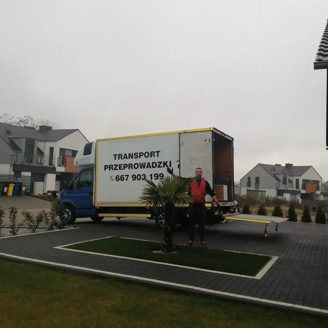 Niebieski bus z otwartą paką i napisem 'TRANSPORT PRZEPROWADZKI' oraz numerem telefonu, zaparkowany na podjeździe nowoczesnego domu, mężczyzna w kamizelce odblaskowej stoi przed busem z rozłożonymi...