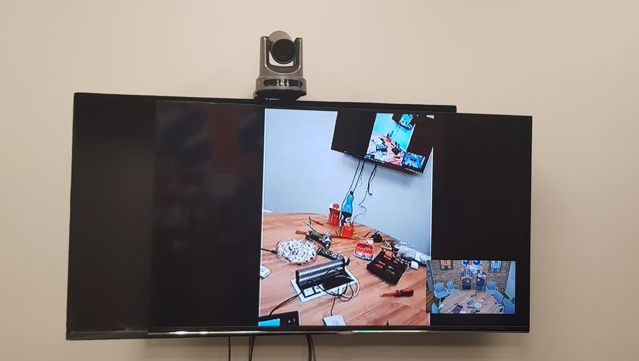 Ekran telewizora z widoczną kamerą wideokonferencyjną, prezentujący obraz stołu roboczego z narzędziami i materiałami elektrycznymi, oraz mniejszy obraz z wideokonferencji.