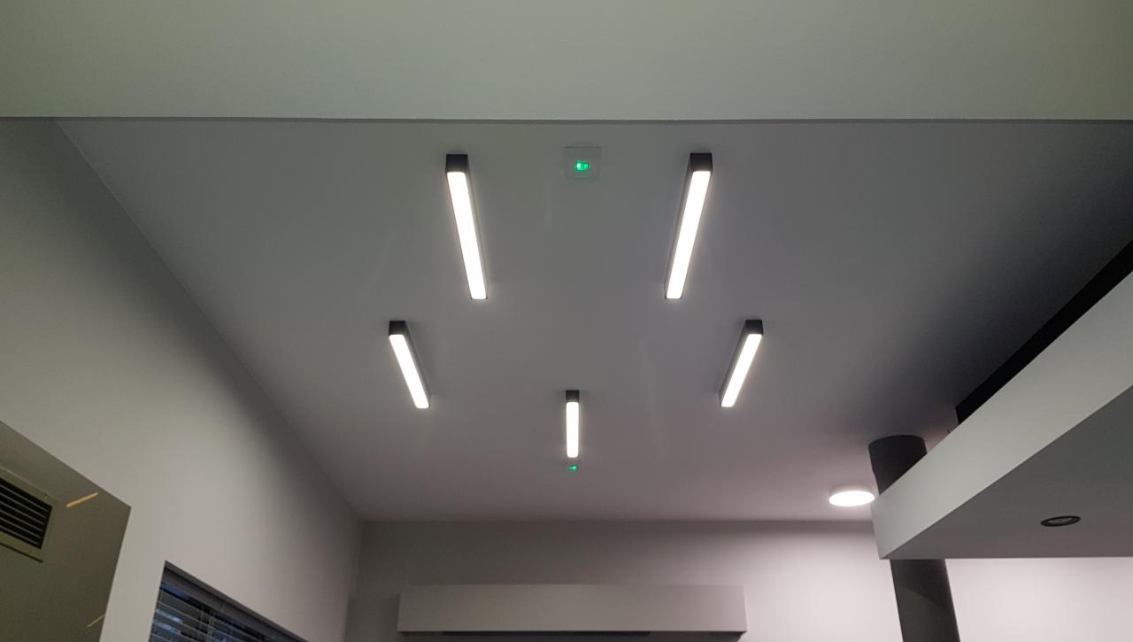 Nowoczesne oświetlenie sufitowe z pięcioma podłużnymi lampami LED z czarnymi obudowami, rozmieszczonymi symetrycznie wokół centralnego punktu z zieloną diodą, na tle gładkiego, szarego sufitu.