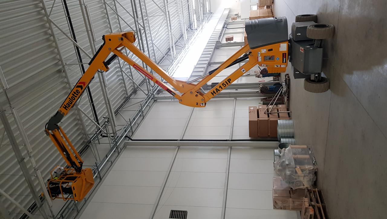 Żółty podnośnik koszowy Haulotte HA15IP w hali magazynowej, widoczne metalowe rury i elementy instalacji pod sufitem, kartony na podłodze.