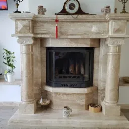 Beżowy kominek z kamienia z czarnym wkładem kominkowym, ozdobiony kolumnami w stylu klasycznym, z detalami dekoracyjnymi na górze, na tle jasnego wnętrza.