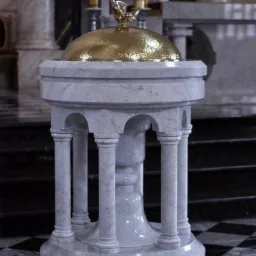 Marmurowa chrzcielnica z kolumnami i złotą kopułą zwieńczoną figurką gołębia, umieszczona w kościele z czarno-białą szachownicą na podłodze.