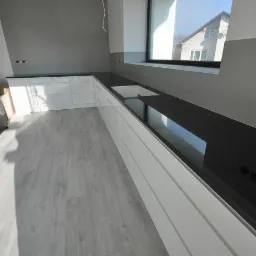 Nowoczesna kuchnia z czarnym blatem, białymi szafkami i szarą podłogą panelową, widok na dom przez okno, widoczne niedokończone gniazdka elektryczne.