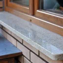 Jasnoszary parapet z konglomeratu zamontowany na ceglanej ścianie pod drewnianym oknem, z dekoracyjną dynią obok.