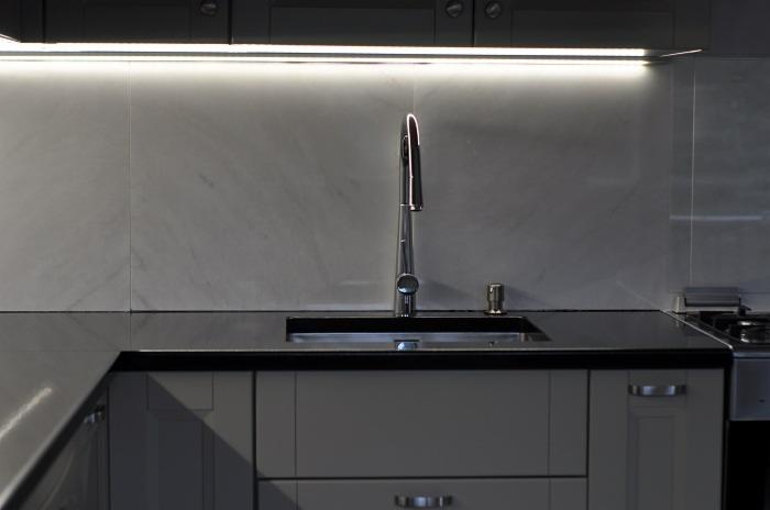 Nowoczesna kuchnia z ciemnym blatem, oświetleniem LED podszafkowym i armaturą ze stali nierdzewnej. Minimalistyczny design z szarymi szafkami i marmurowym panelem ściennym.