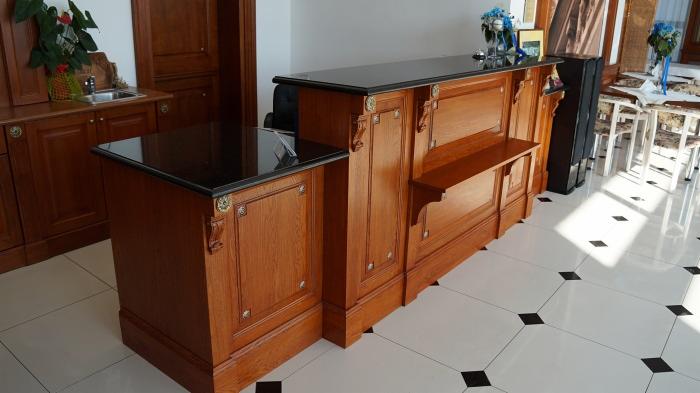 Drewniana lada recepcyjna z czarnym blatem z kamienia, zdobiona rzeźbieniami, na białej podłodze w czarne romby.