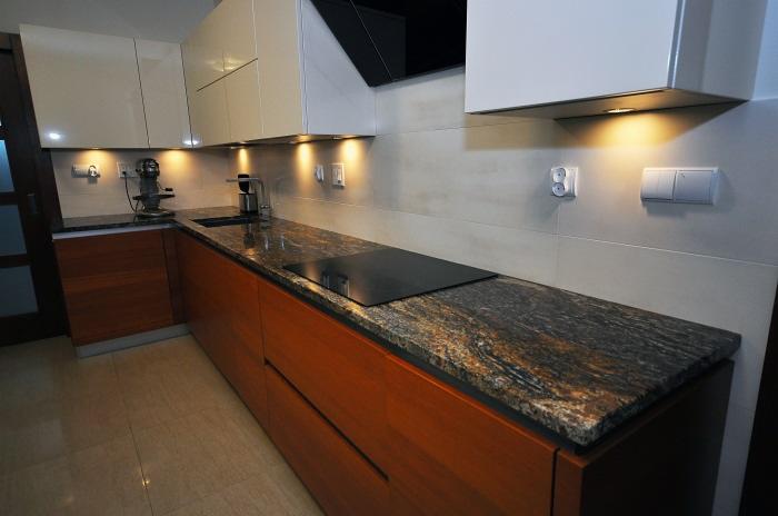 Kuchnia z blatem o unikalnym wzorze kamienia, zabudowanym z płytą grzewczą, oświetleniem podszafkowym i jasnymi szafkami wiszącymi.