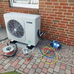 Biała jednostka zewnętrzna pompy ciepła marki York, podłączona do manometru z kolorowymi przewodami, stoi na kostce brukowej przed ceglaną ścianą budynku z oknem.