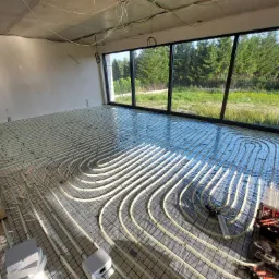 Wnętrze w trakcie instalacji ogrzewania podłogowego, widoczne rury grzewcze ułożone na siatce, narzędzia i materiały budowlane obok, duże okno z widokiem na zieleń na zewnątrz.