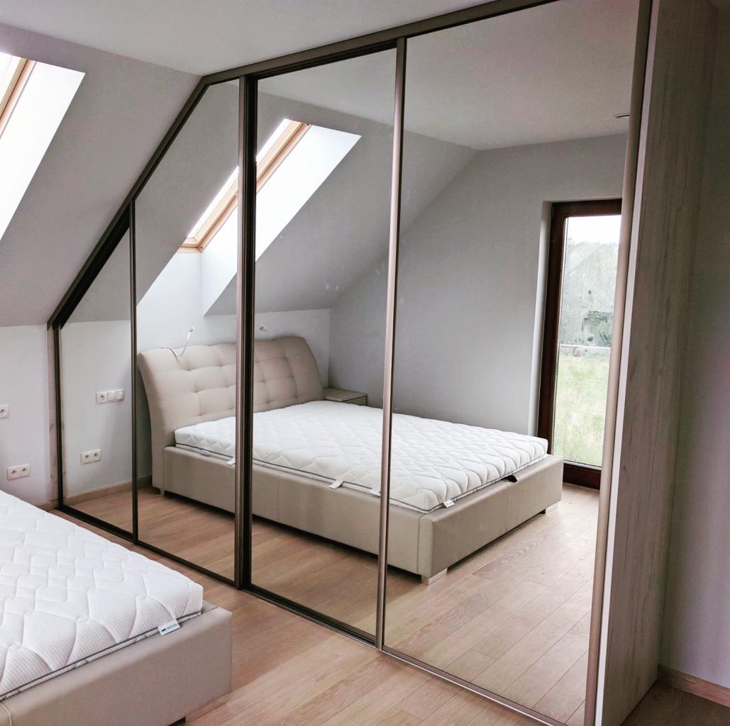 Szafa przesuwna z lustrzanymi drzwiami w pokoju na poddaszu, odbicie łóżka z pikowanym zagłówkiem i okna dachowe.