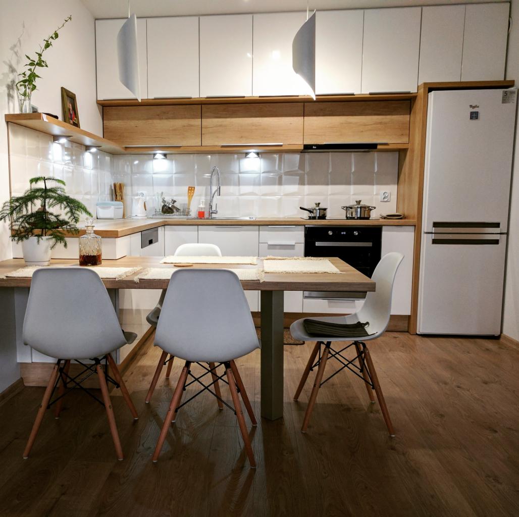 Jasna kuchnia z białymi frontami szafek i drewnianymi akcentami, stół z szarymi krzesłami w stylu skandynawskim, na blacie kuchennym doniczka z iglakiem i szklana karafka