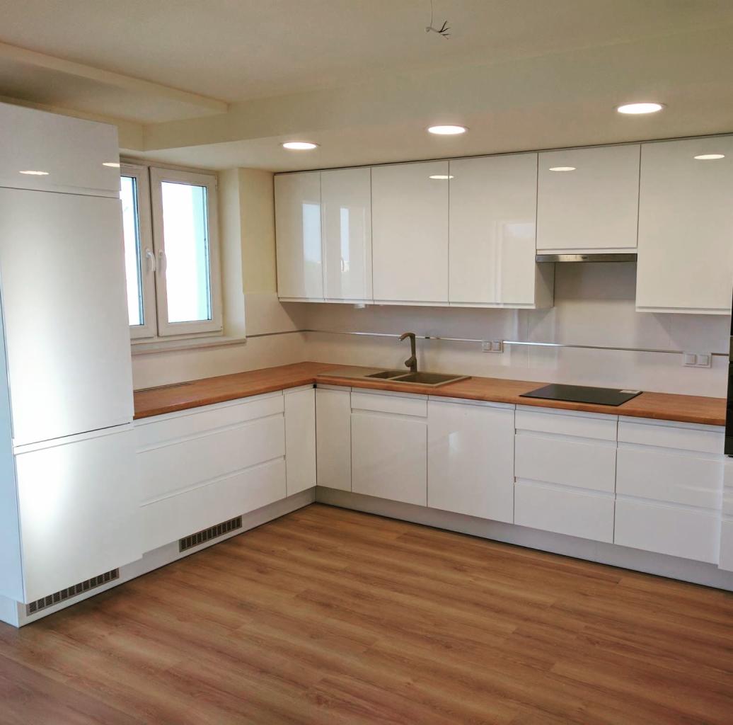 Biała, nowoczesna kuchnia z drewnianym blatem, oświetleniem punktowym i brązową podłogą z paneli, widok na okno i część zabudowy kuchennej.