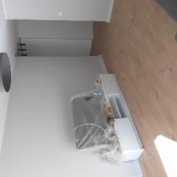 Wnętrze pokoju w trakcie remontu, widoczna biała szafka RTV pod folią ochronną, szara lampa sufitowa, panele podłogowe i białe ściany.