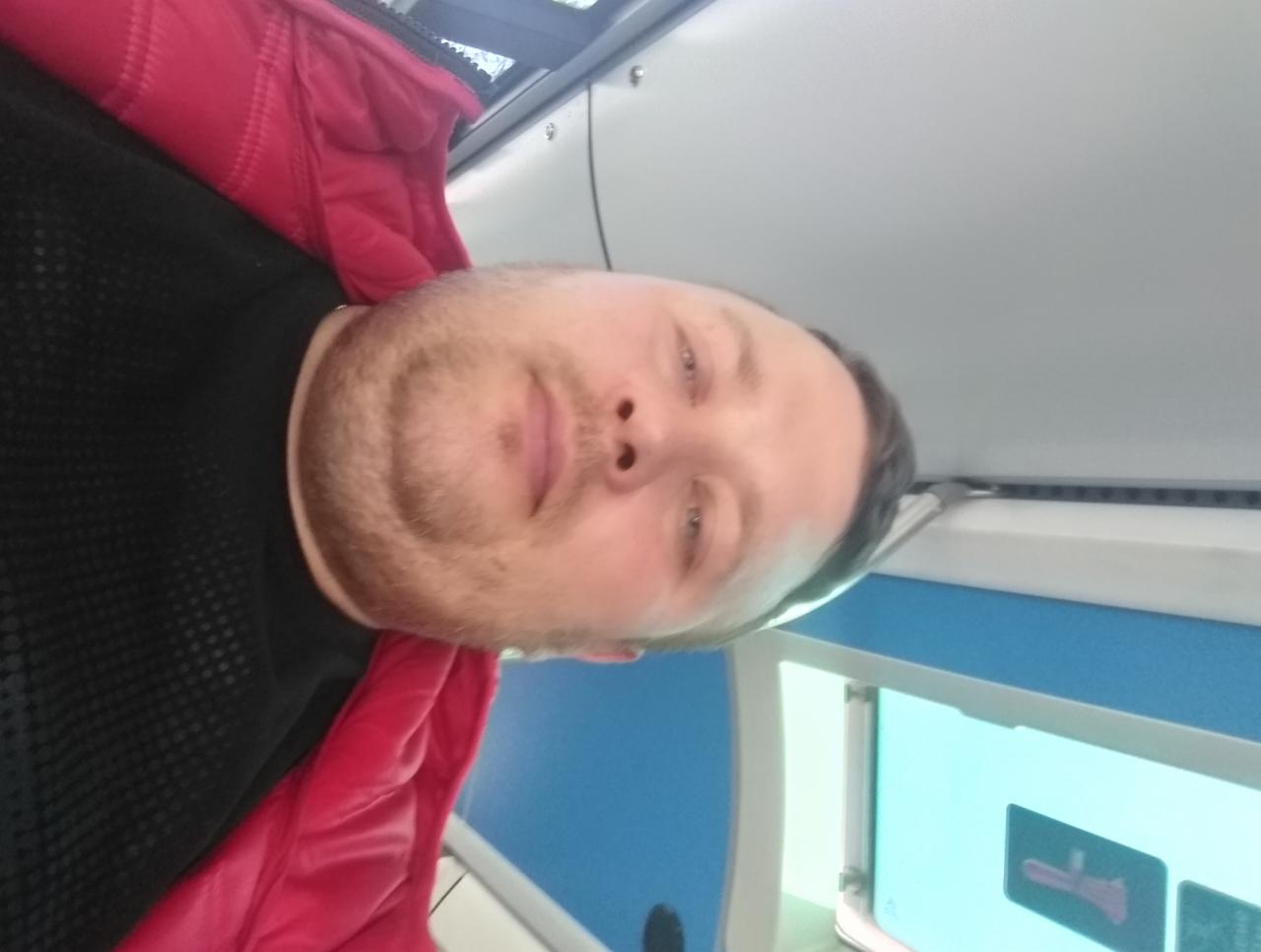 Portret mężczyzny w czerwonej kurtce na tle wnętrza wagonu kolejowego, z widocznym oknem i naklejką informacyjną. Zdjęcie obrócone o 90 stopni.