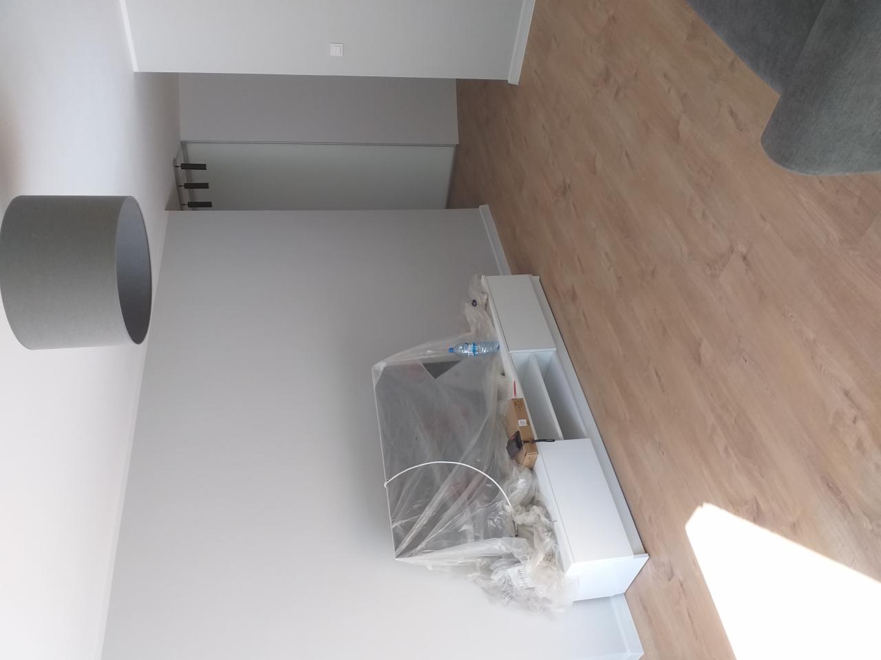 Wnętrze pokoju w trakcie remontu, widoczna biała szafka RTV pod folią ochronną, szara lampa sufitowa, panele podłogowe i białe ściany.