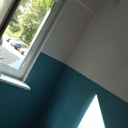 Świeżo pomalowane wnętrze pokoju na biało i turkusowo, z otwartym oknem wpuszczającym jasne światło słoneczne, widoczna drewniana podłoga i gniazdko elektryczne.