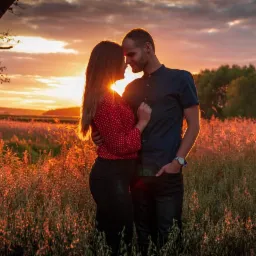 Zakochana para w objęciach stoi na tle zachodzącego słońca w polu wysokich traw, sylwetki drzew po lewej stronie kadru, ciepłe światło.