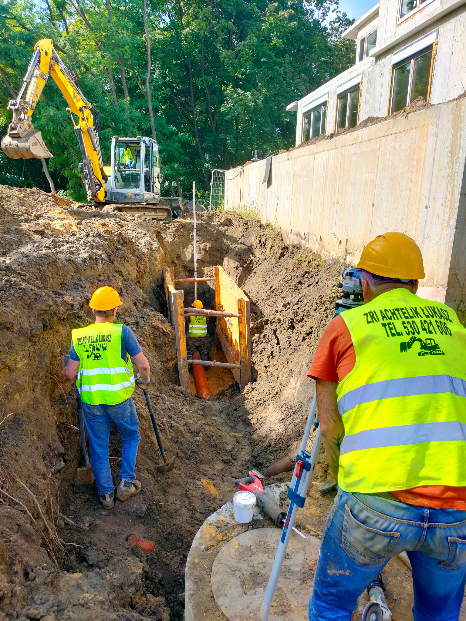 Prace ziemne: koparka wykopuje rów, robotnicy w żółtych kamizelkach i kaskach układają rury w zabezpieczonym wykopie, geodeta mierzy poziom terenu.