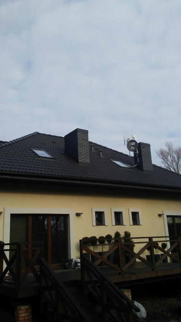 Dom z ciemną dachówką, dwoma kominami, oknami dachowymi i anteną satelitarną