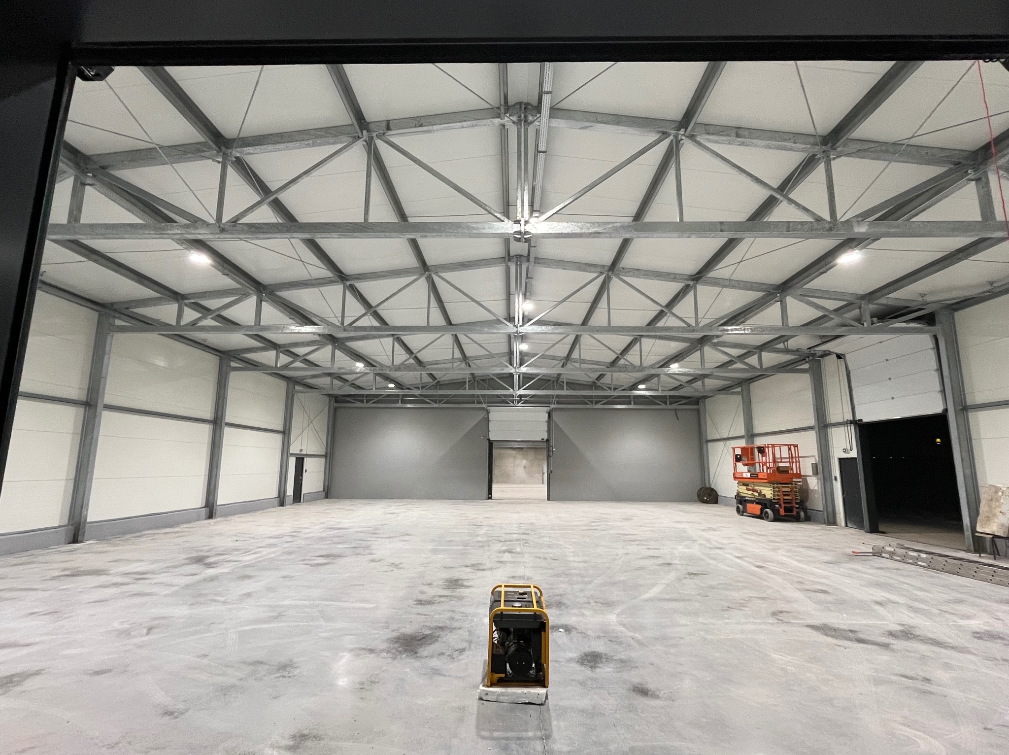 Wnętrze hali magazynowej z widoczną konstrukcją stalową dachu, oświetleniem LED, podnośnikiem koszowym w tle i agregatem prądotwórczym na pierwszym planie.