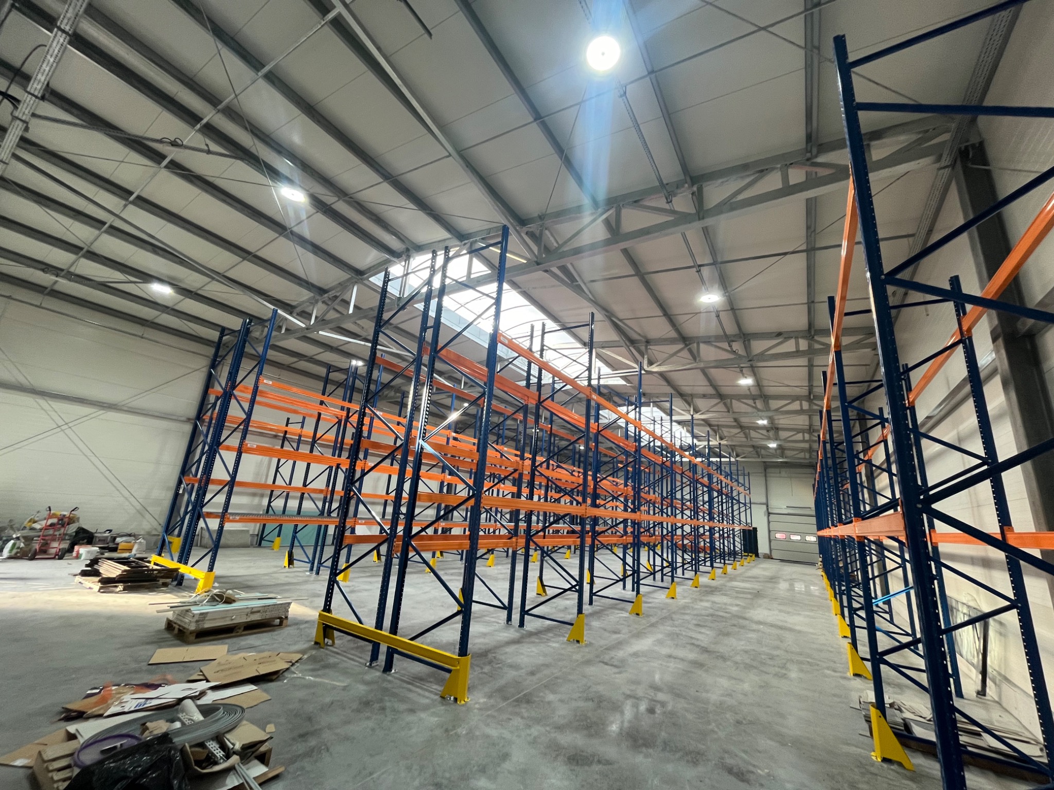Wnętrze hali magazynowej z zamontowanymi, niezapełnionymi regałami metalowymi w kolorach niebieskim i pomarańczowym, widoczne oświetlenie sufitowe oraz resztki materiałów budowlanych na podłodze.