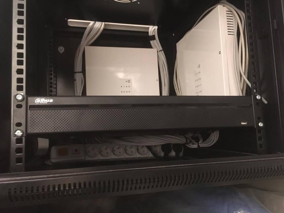Szafa rack z zamontowanym rejestratorem Dahua, dwoma białymi centralami i listwą zasilającą; widoczne okablowanie.