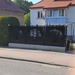 TOMASZ JASKÓLSKI - Nowoczesne, czarne ogrodzenie panelowe z motywem drzewa, oddzielające posesję od chodnika. Widoczne dwa budynki w tle i fragment zieleni.