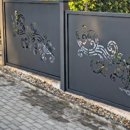 TOMASZ JASKÓLSKI - Nowoczesne, ciemnoszare przęsła metalowego ogrodzenia z laserowo wycinanymi, falistymi wzorami roślinnymi, tworzące bramę przesuwną na podjeździe z kostki brukowej w Bodzanowie. Widoczne kamyki...