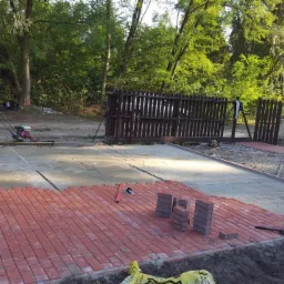 Częściowo ułożona czerwona kostka brukowa na przygotowanym podłożu, widoczne narzędzia, maszyna do zagęszczania gruntu i samochód w tle.