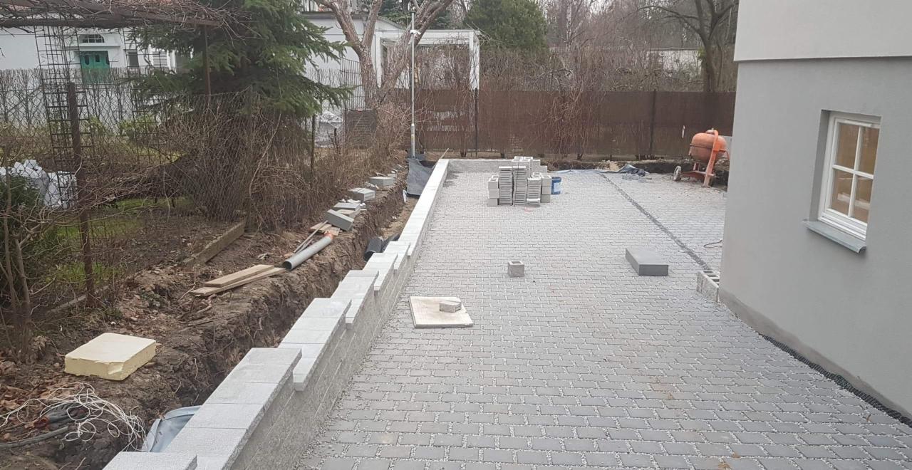 Wykładanie szarej kostki brukowej na podwórku, widoczny fragment murku oporowego w trakcie budowy, w tle betoniarka i budynek z szarą elewacją.