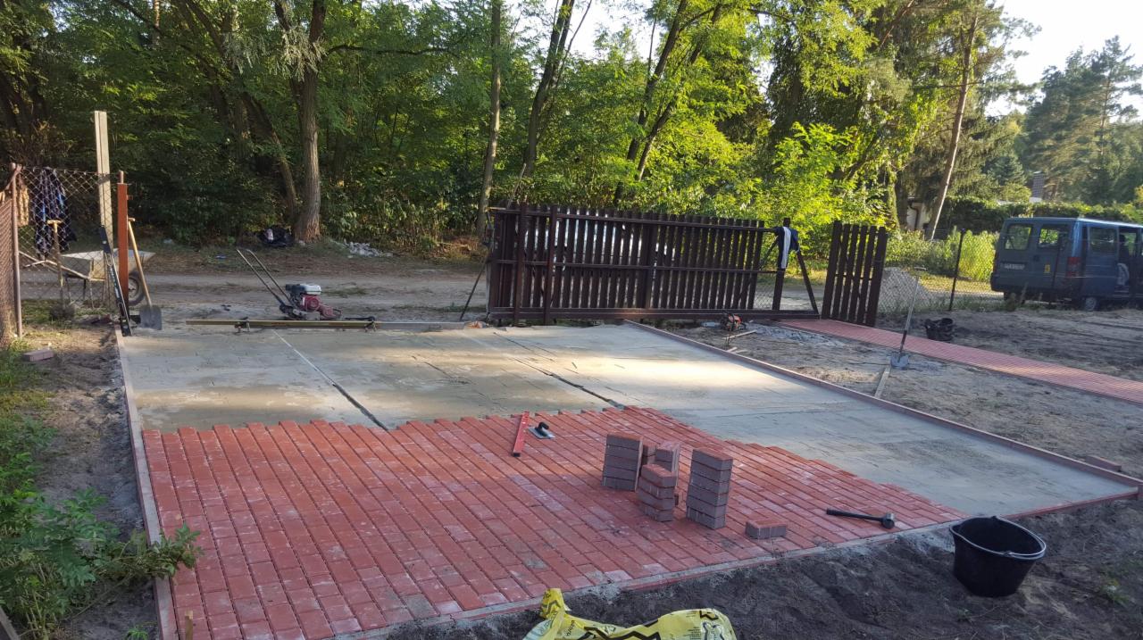 Częściowo ułożona czerwona kostka brukowa na przygotowanym podłożu, widoczne narzędzia, maszyna do zagęszczania gruntu i samochód w tle.