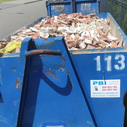 PBI Spółka Jawna - Niebieski kontener na gruz, wypełniony potłuczonymi cegłami i fragmentami płytek, z widocznym logo firmy PBI Częstochowa i numerem 113, ustawiony na trawie przy chodniku.