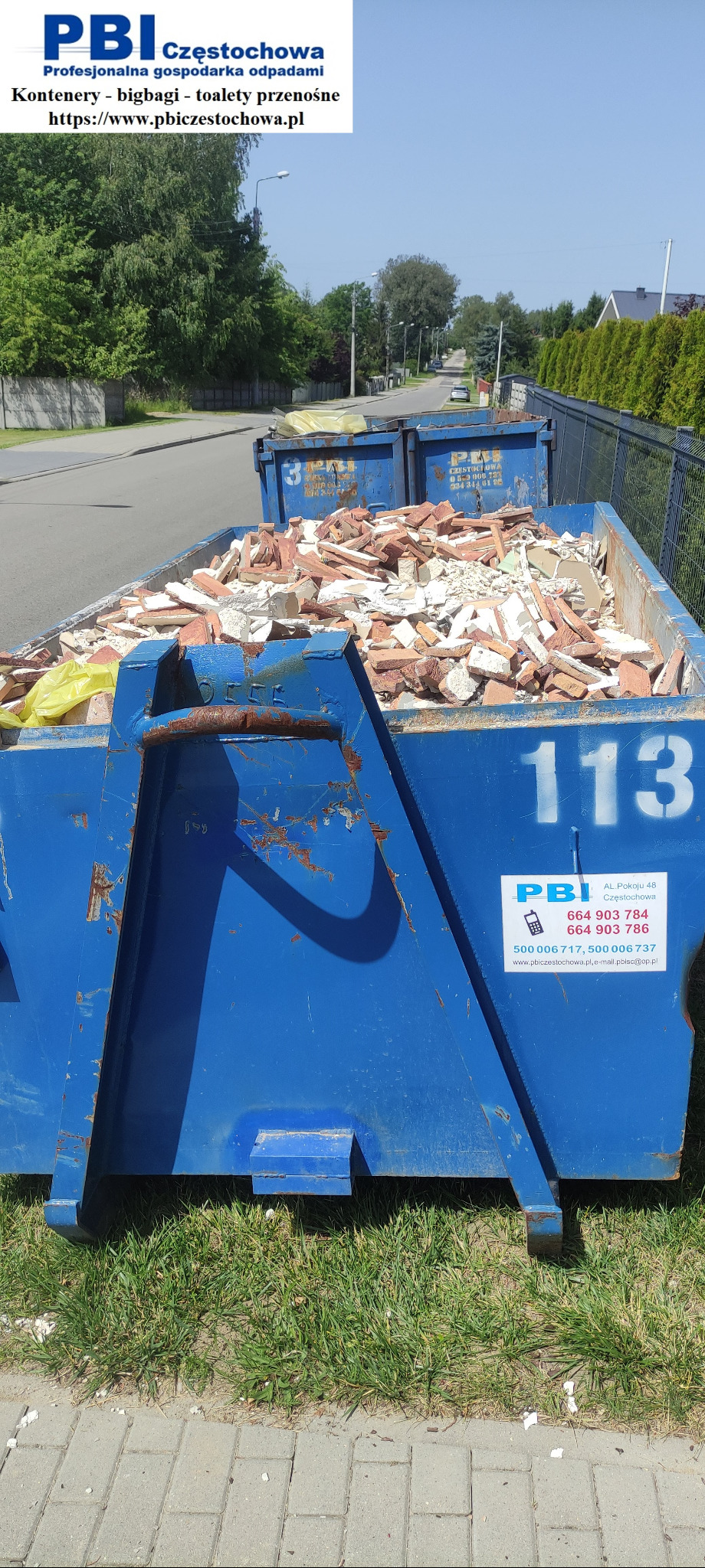 Niebieski kontener na gruz, wypełniony potłuczonymi cegłami i fragmentami płytek, z widocznym logo firmy PBI Częstochowa i numerem 113, ustawiony na trawie przy chodniku.