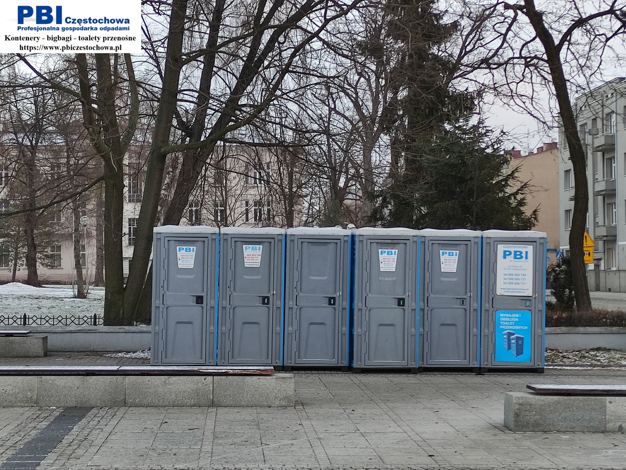Szereg szarych, przenośnych toalet z niebieskimi akcentami, ustawionych na chodniku w miejskim otoczeniu, z widocznymi budynkami w tle i drzewami bez liści, sugerującymi porę zimową.