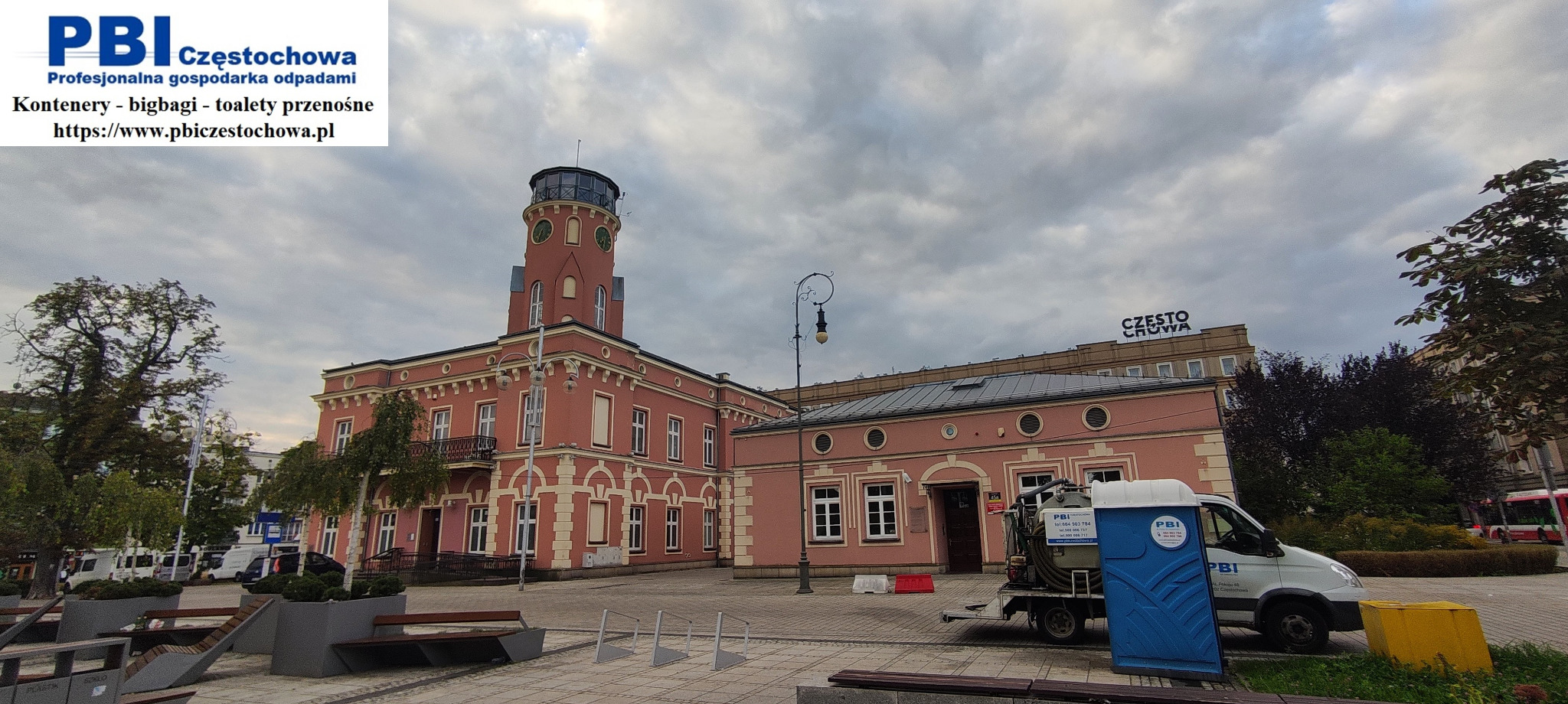 Przenośna toaleta na przyczepce zaparkowana na placu miejskim przed budynkiem z wieżą zegarową w Częstochowie.