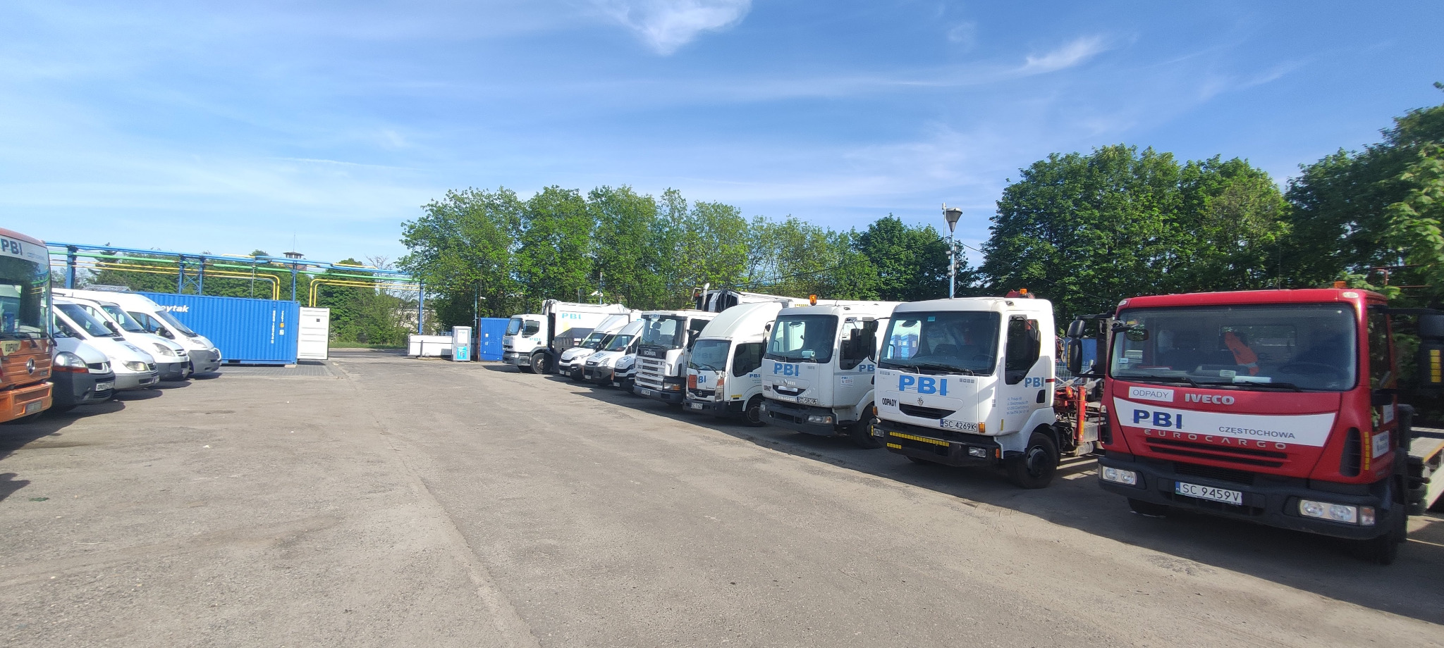 Rząd pojazdów firmowych PBI, w tym ciężarówki i busy, zaparkowanych na placu w słoneczny dzień. Widoczny niebieski kontener i rurociągi w tle.
