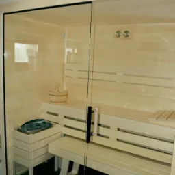 Jasna, drewniana kabina z przeszklonymi drzwiami, widoczne ławki, wiadro i piec na kamienie, oświetlenie punktowe na suficie.