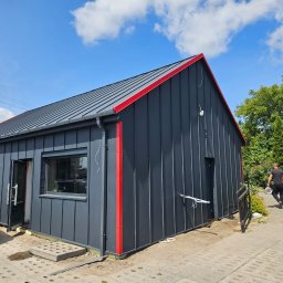 Dachy/Ciesielstwo - Nowoczesny budynek z grafitową blachą na elewacji i dachu, z czerwonymi akcentami. Widoczny fragment samochodu i osoba z narzędziami w tle.