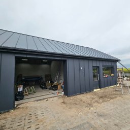 Dachy/Ciesielstwo - Szary budynek z blachy trapezowej z nowym dachem z blachy na rąbek stojący. Widoczna drabina i narzędzia w otwartym garażu. Trawa w tle.