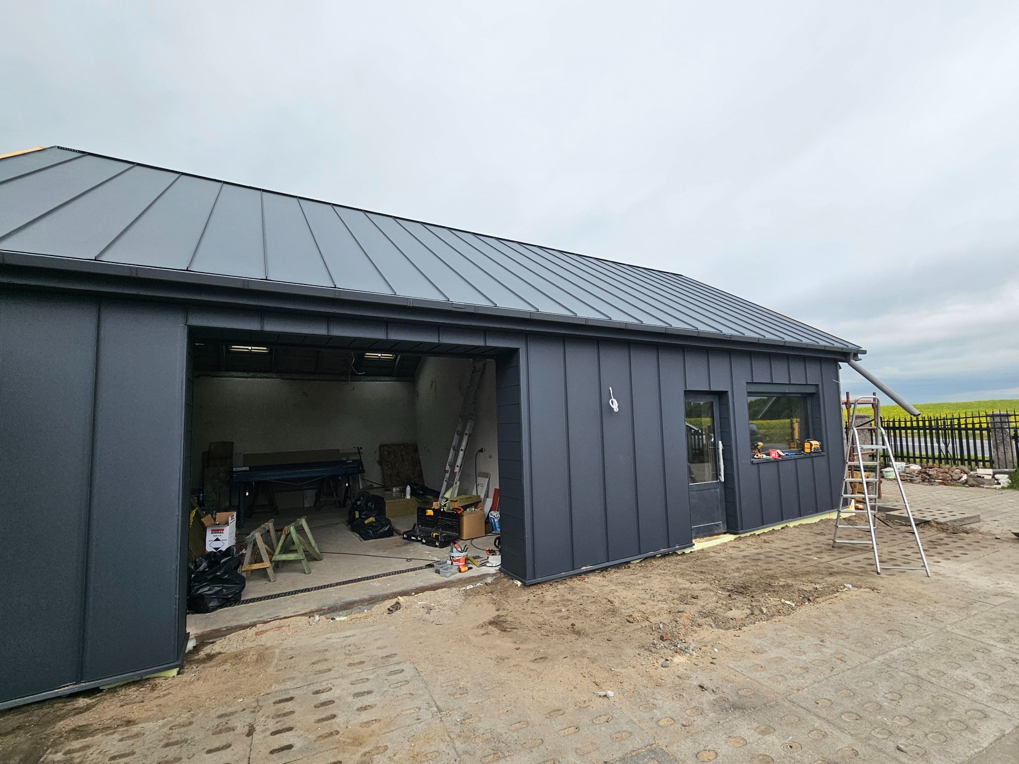 Nowoczesny, grafitowy budynek gospodarczy z blachy trapezowej. Widoczny fragment dachu z rynnami, okno i drzwi. Na zewnątrz drabina i narzędzia.