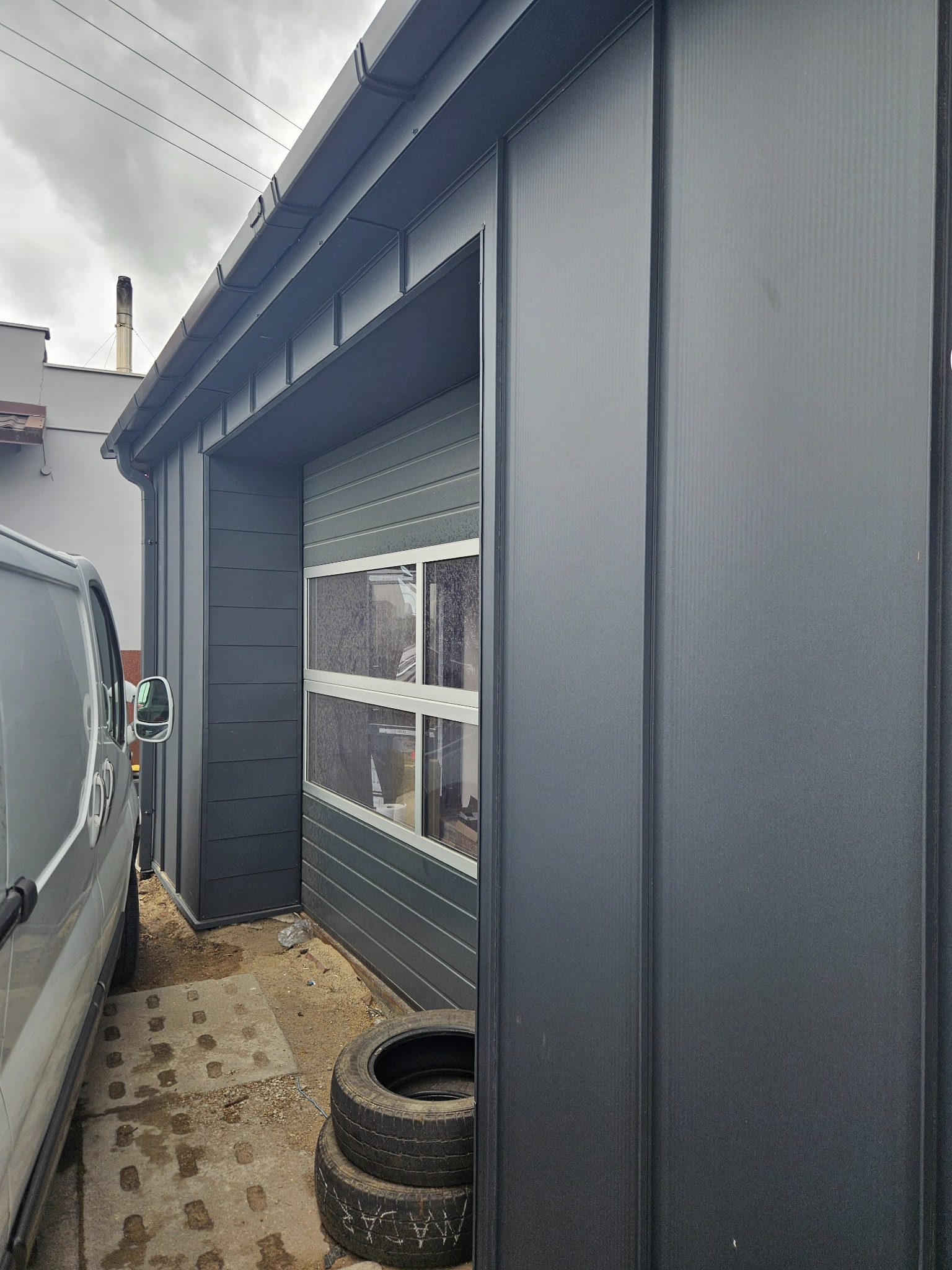 Szary garaż blaszany z segmentową bramą i rynnami, widoczny z boku. Obok zaparkowany samochód dostawczy i opony. Pochmurne niebo w tle.