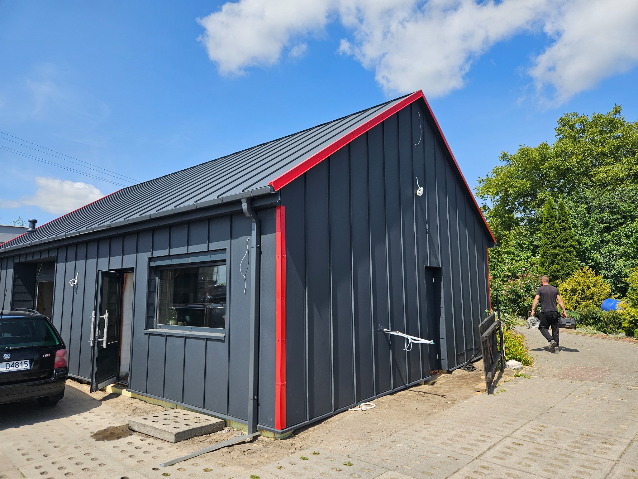 Nowoczesny budynek z grafitową blachą na elewacji i dachu, z czerwonymi akcentami. Widoczny fragment samochodu i osoba z narzędziami w tle.