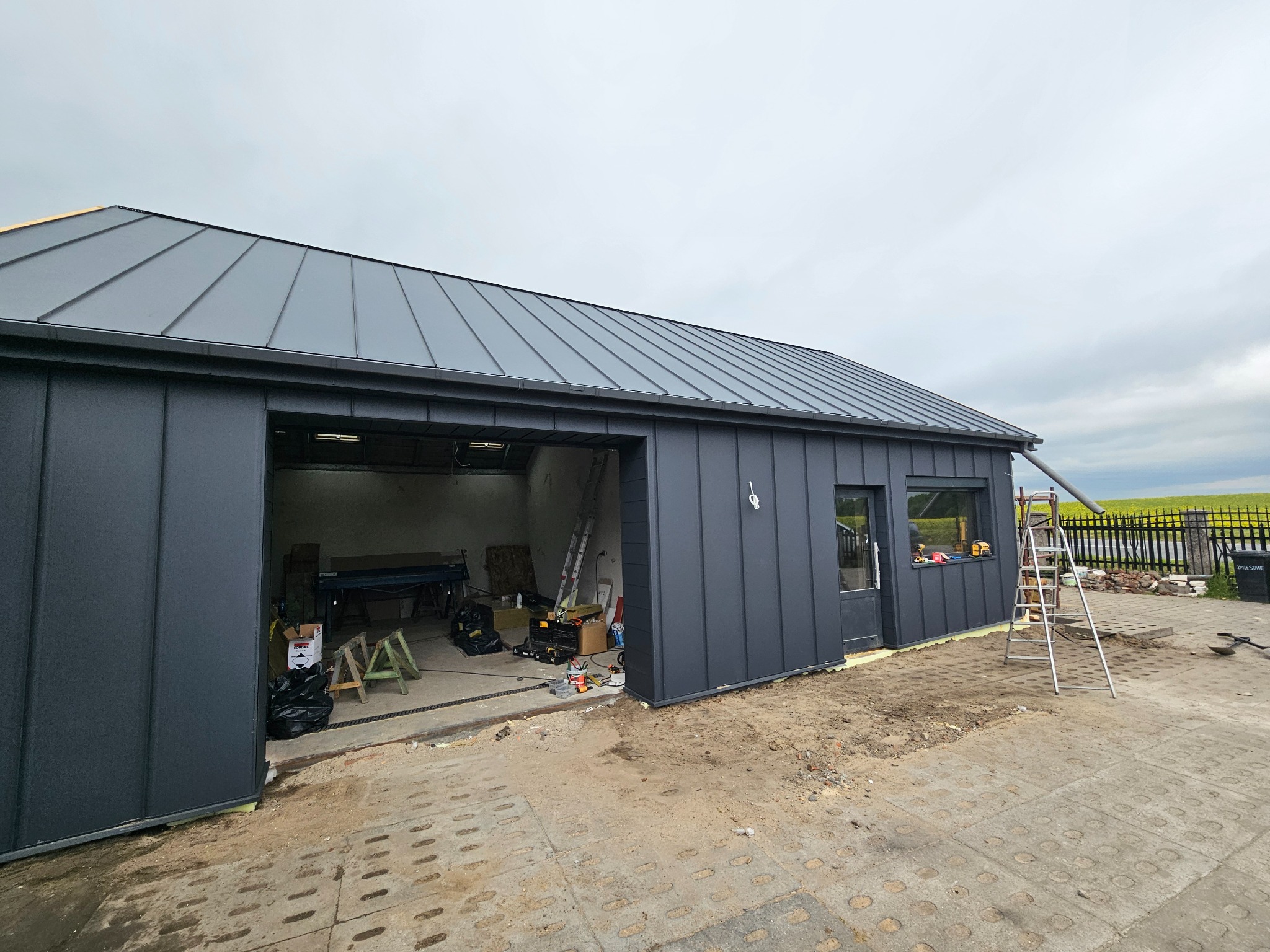 Szary budynek z blachy trapezowej z nowym dachem z blachy na rąbek stojący. Widoczna drabina i narzędzia w otwartym garażu. Trawa w tle.